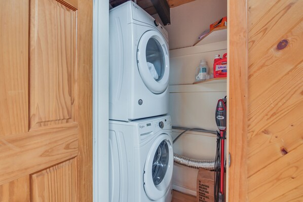 Une petite buanderie équipée d’un lave-linge et d’un sèche-linge à chargement frontal, d’une étagère pour les produits de nettoyage et d’un tuyau pour les raccordements à l’eau.
