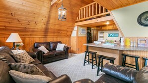 Un acogedor interior de cabaña con techo de madera, una sala de estar con sofás de cuero, una zona de cocina con barra, y un reloj a la vista.