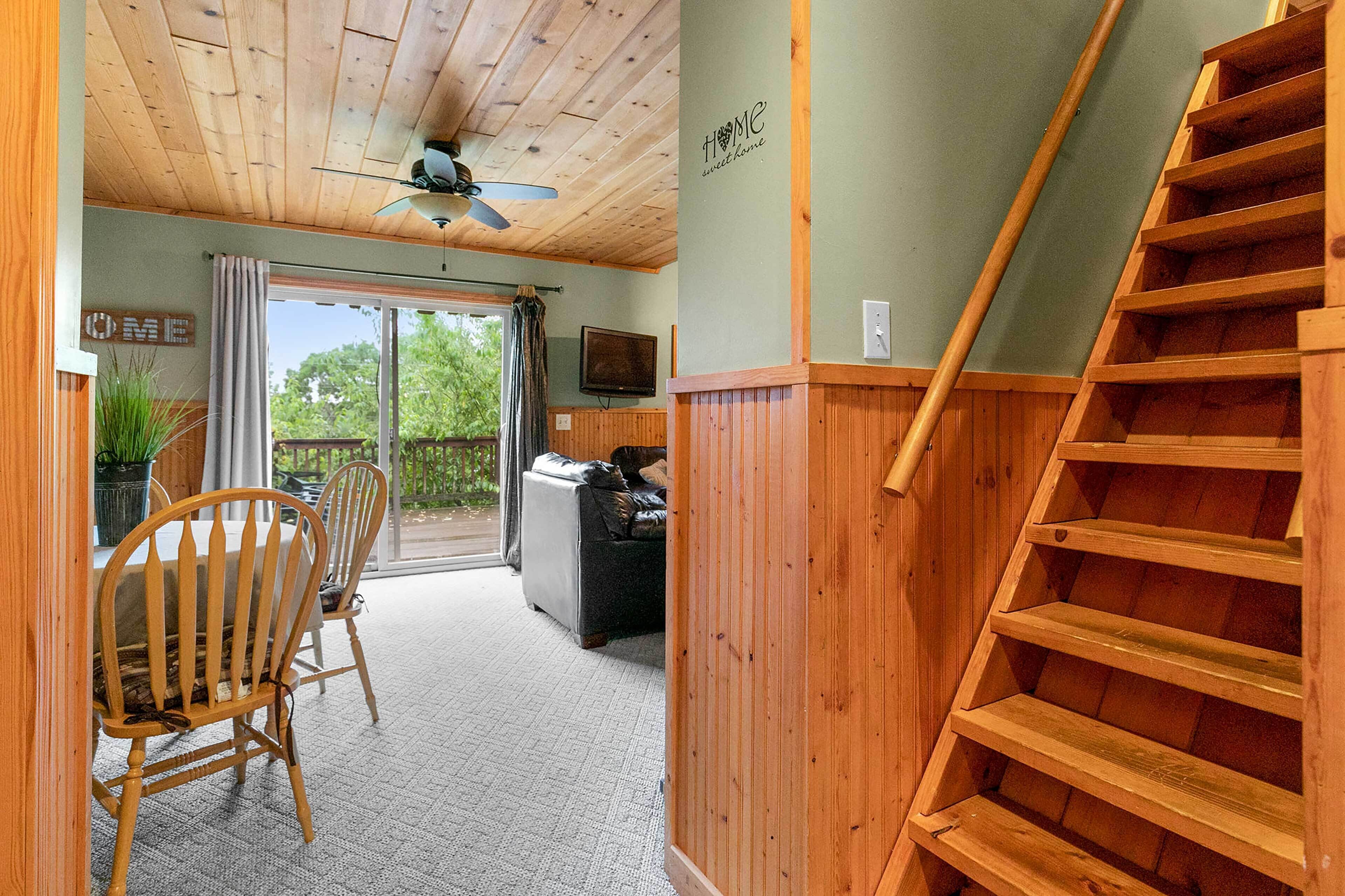 A wooden staircase leading to a balcony, a dining area with chairs and a table, and a television on the wall.