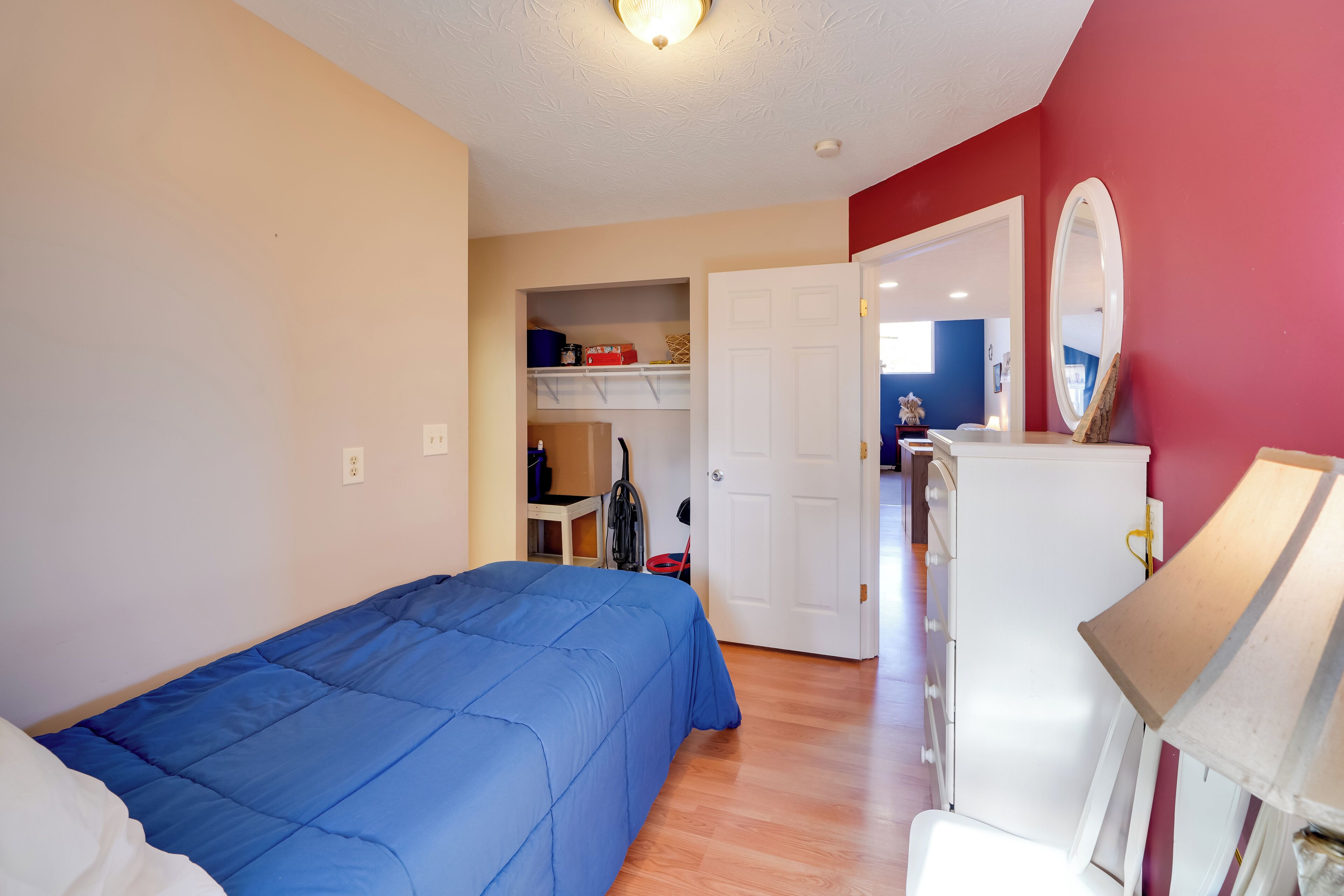 A bedroom with a blue bedspread, a white dresser, a mirror, and a closet with shelves.