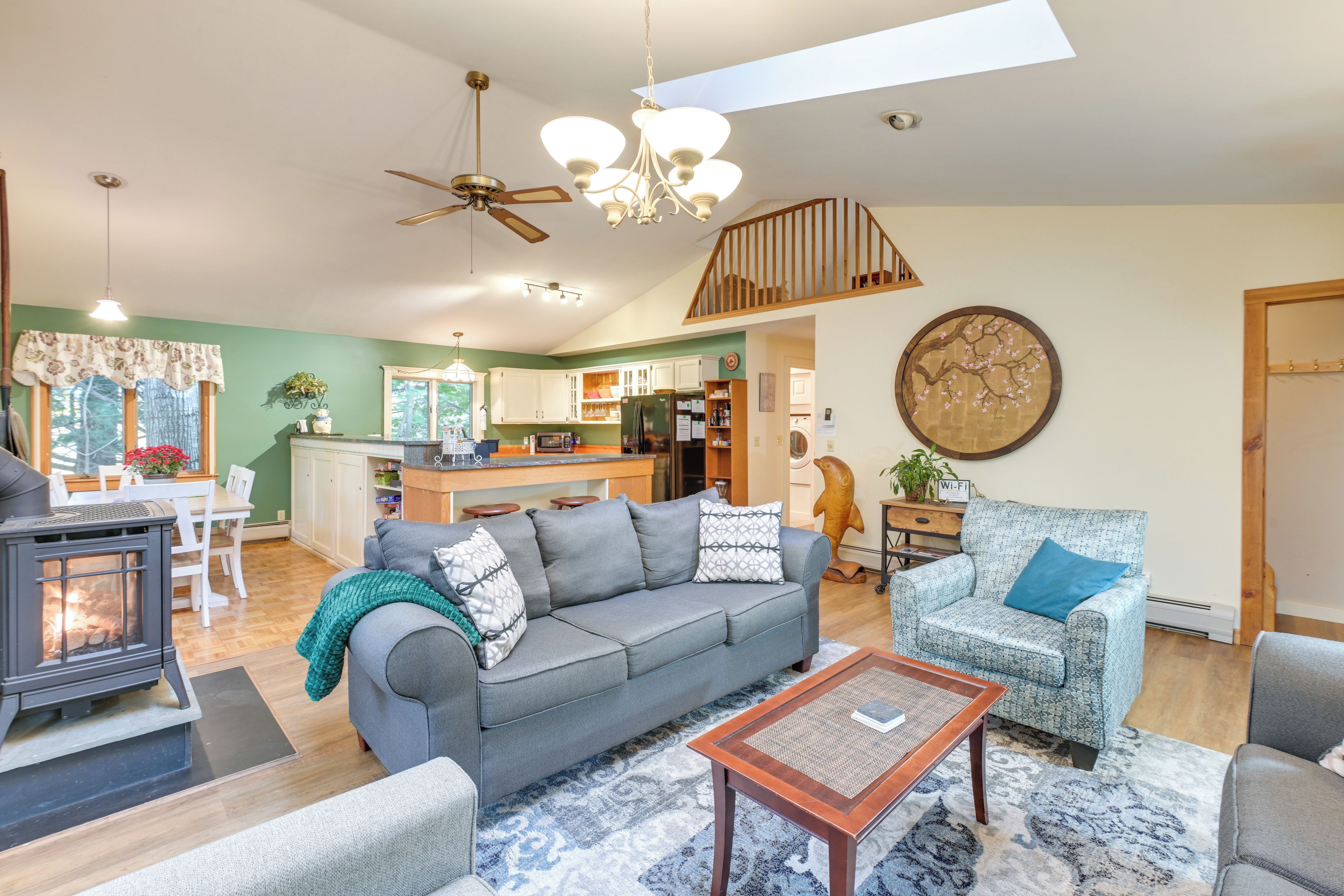 A spacious living area with a sofa, armchairs, and a coffee table. A fireplace is visible in the foreground, and a kitchen is in the background.