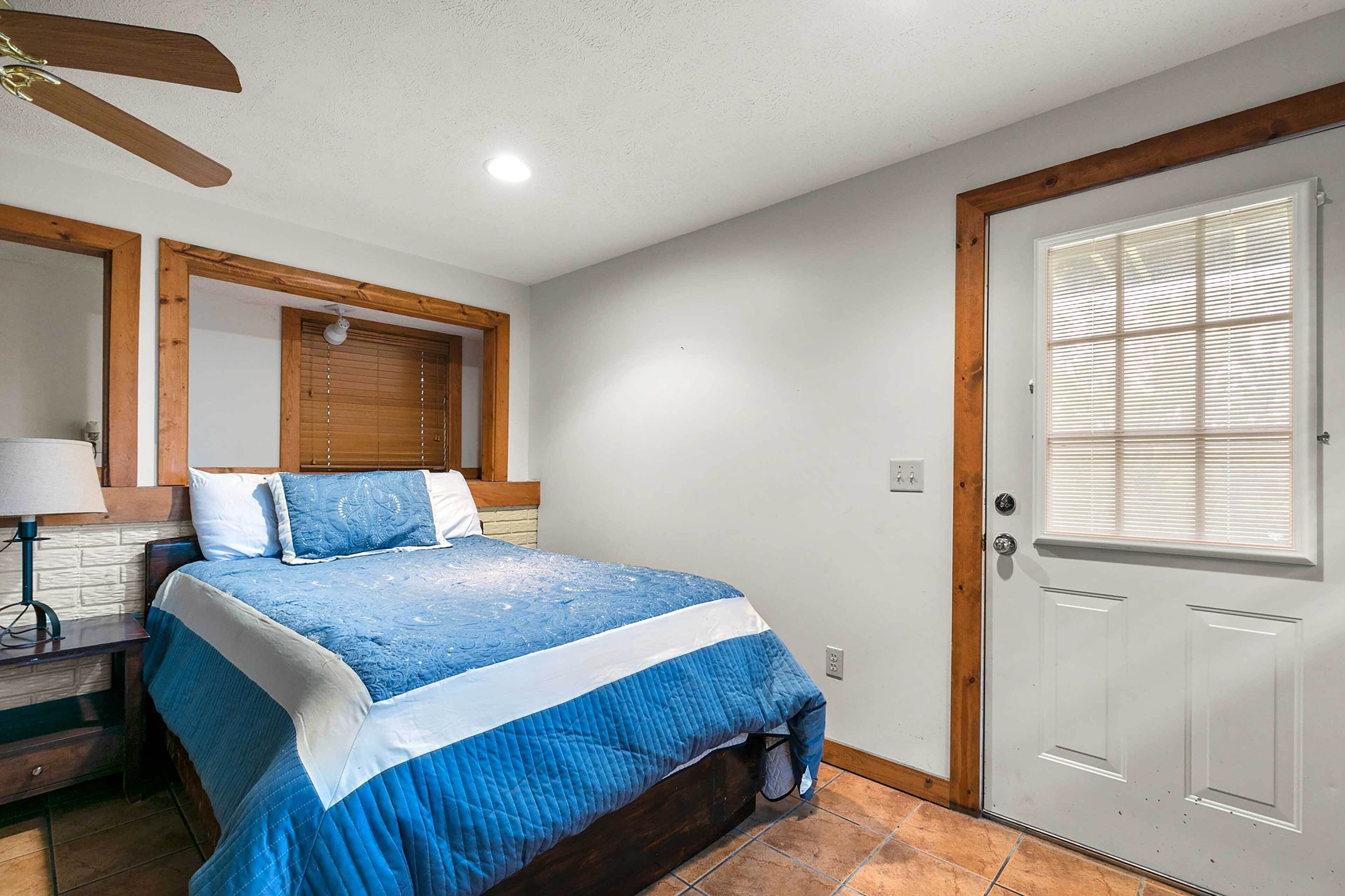 Une chambre à coucher avec un lit, une tête de lit en bois, une porte avec des stores, une table de chevet avec une lampe et un ventilateur de plafond.