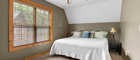 Un dormitorio con una cama, un ventilador de techo, una ventana con persianas, una mesita de noche con lámpara y un anclaje de pared.