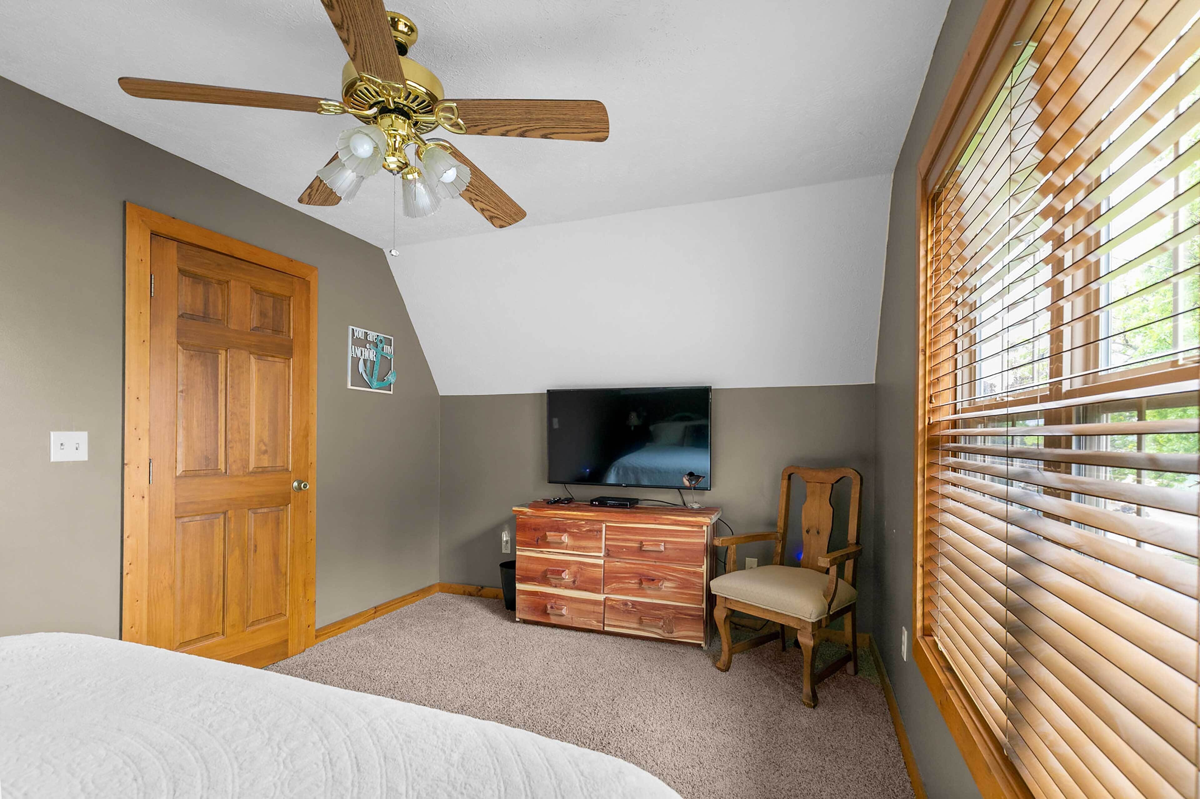 A bedroom with a wooden door, a ceiling fan, a TV, a dresser, a chair, and a window with blinds.