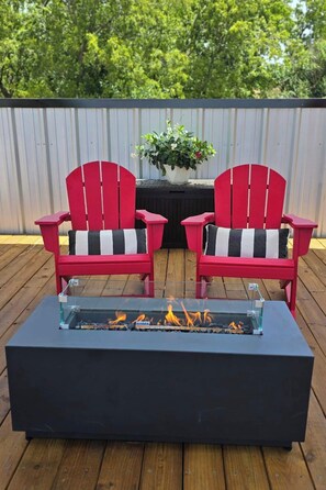 Two red Adirondack chairs with striped cushions on a wooden deck, a glass-top fire pit in the center, and a potted plant on a table in the background. - Historic Stay in Santa Fe Railcar (Brownwood)