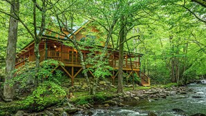 A wooden cabin with a deck and stairs, surrounded by lush greenery and a flowing river. - Beautiful Three Bedroom Cabin Directly On Cosby Creek (Cosby)