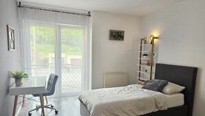A bedroom with a bed, a desk, a chair, a radiator, and a window with curtains. - Apartment on the edge of the Palatinate Forest. TLA/TDY (Ramstein-Miesenbach)