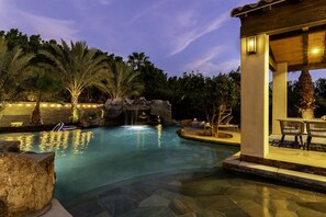 A swimming pool with a waterfall feature, surrounded by palm trees and a stone-tiled deck.