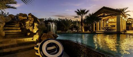 A pool area with a waterfall, palm trees, and a building with outdoor seating.