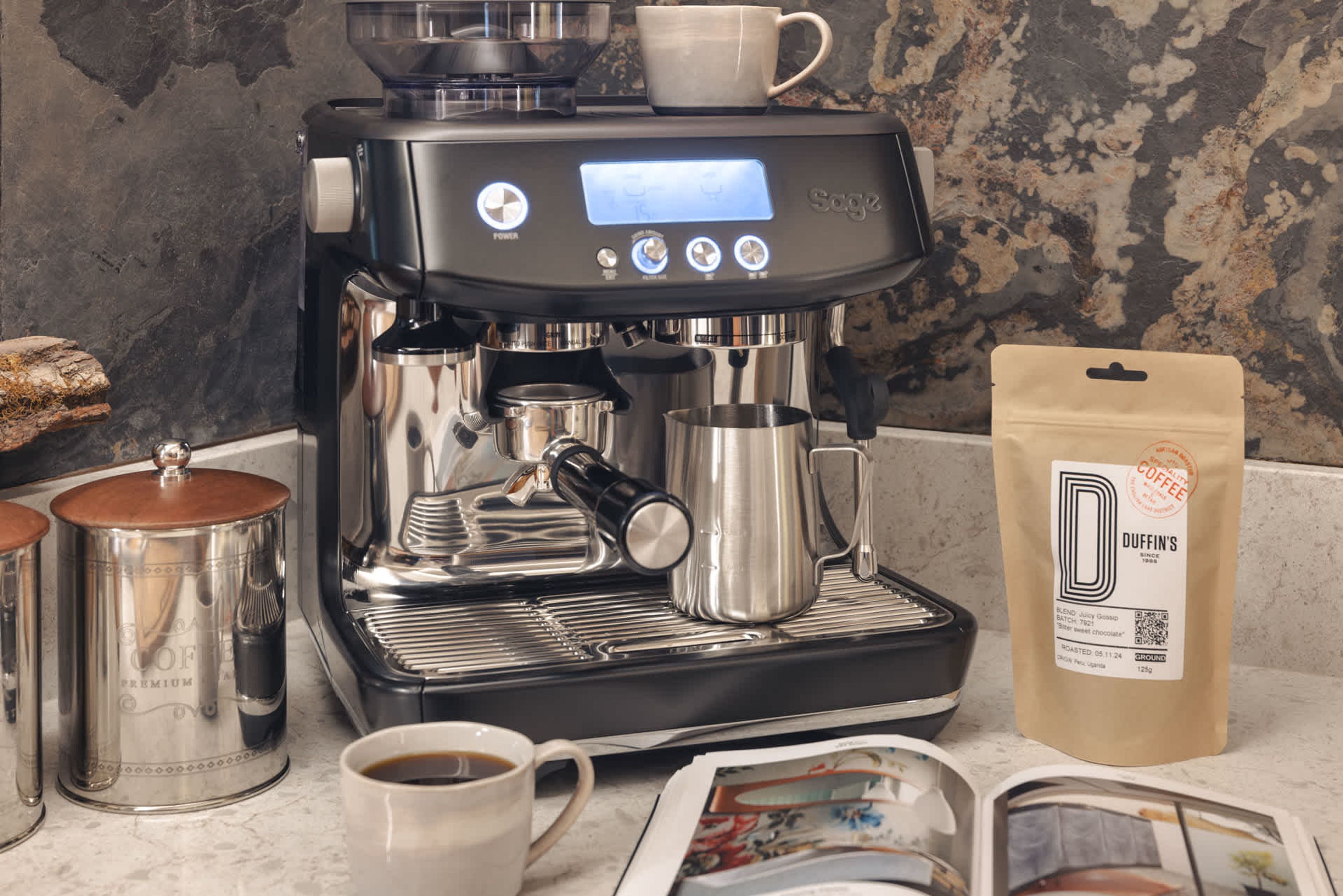 A coffee machine with a digital display, a coffee cup, a bag of coffee, and two stainless steel containers.