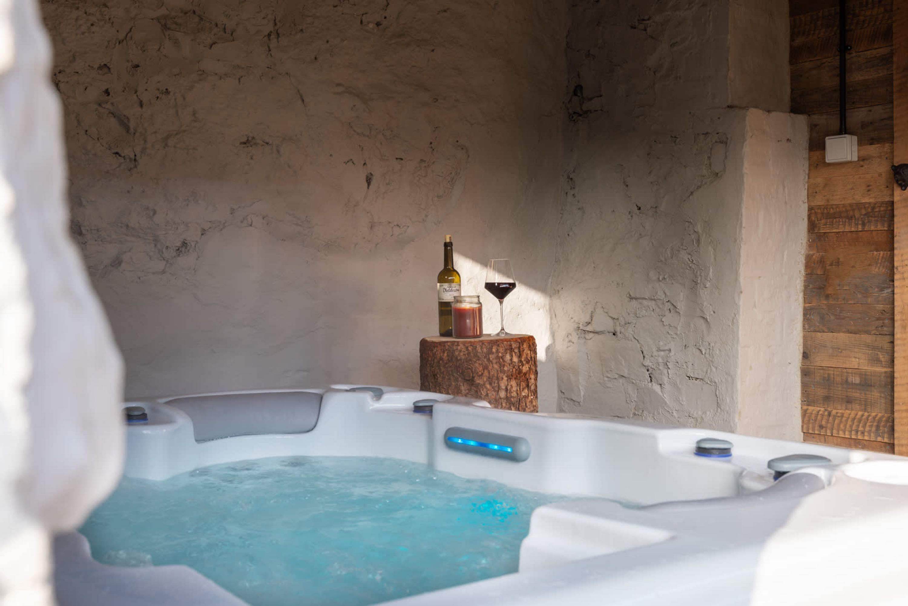 A hot tub with a wooden stool beside it, a bottle, and a glass of red wine.