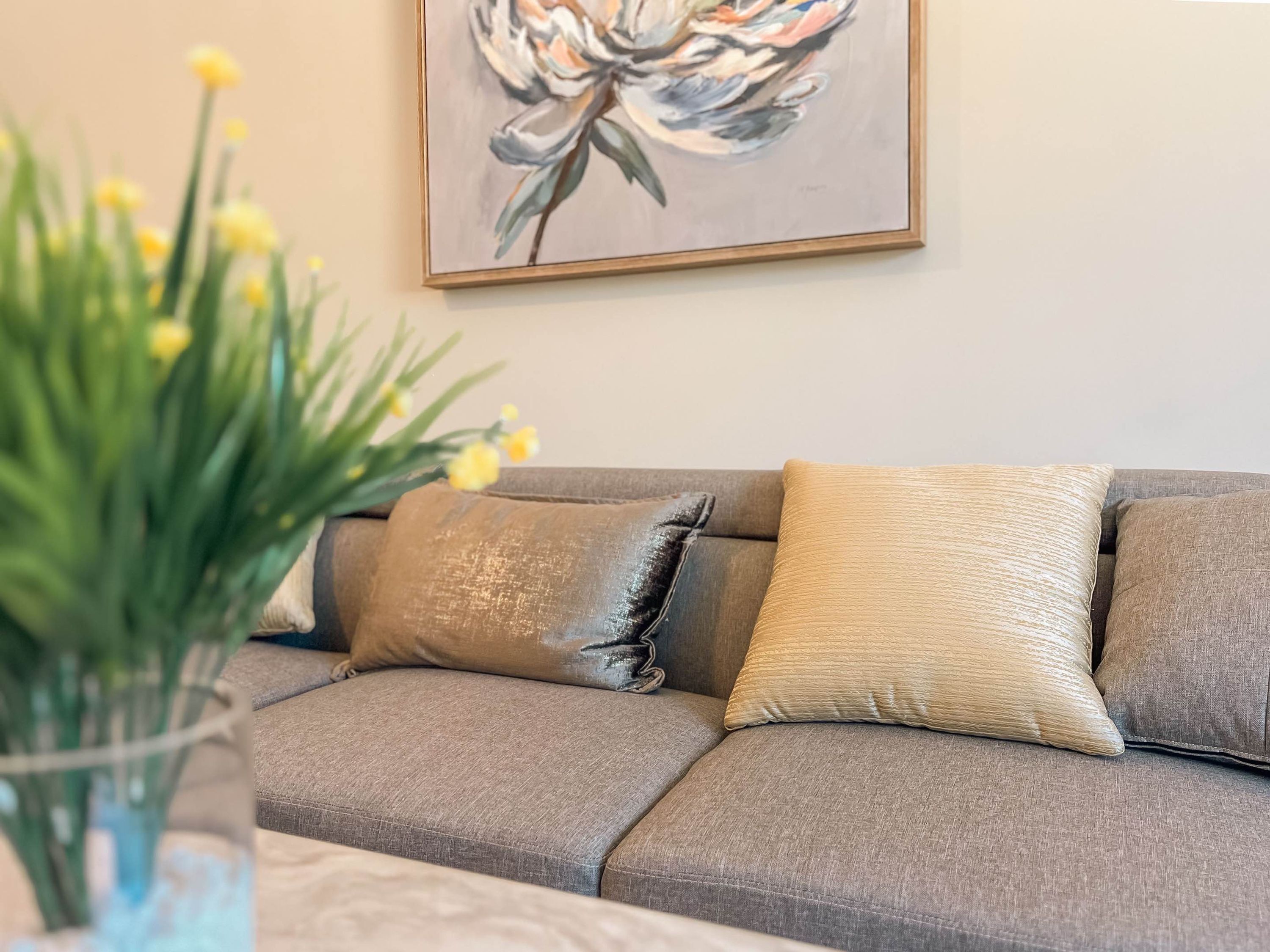 A living room with a grey sofa, a vase with yellow flowers, and a painting on the wall.