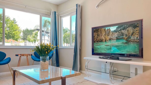 A modern living room with a glass-top coffee table, a flat-screen TV on a white stand, blue chairs, and a view of a lake through the window. - Modern Retreat With Spacious Yard Cozy Interiors (Ontario)