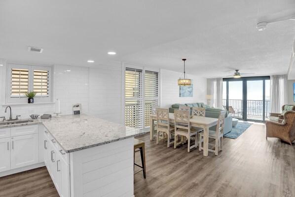 A modern kitchen with white cabinetry, a marble countertop, and a dining area with a wooden table and chairs. - Wind Drift 401 by Liquid Life (Orange Beach)