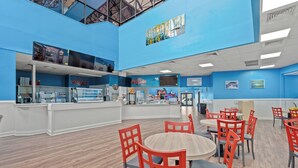 A spacious indoor area with blue walls, a counter with a pizza display, and multiple seating areas with red chairs. - Spacious Beachfront Condo at Sand Dunes Resort 1203 – Perfect Location with Top (Myrtle Beach)