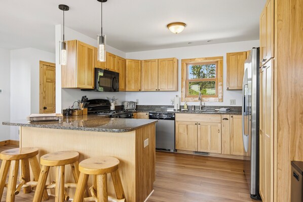 A modern kitchen with wooden cabinets, a central island with stools, stainless steel appliances, and a window providing natural light.