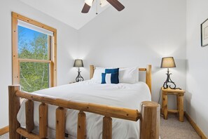 Un dormitorio con estructura de cama de madera, dos mesitas de noche con lámparas, una ventana con vista a árboles y un ventilador de techo.