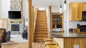 A modern interior with a stone fireplace, wooden staircase, kitchen area, and seating space.