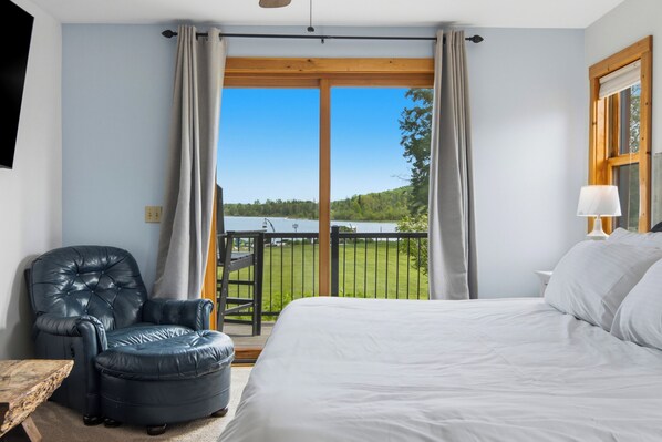 A bedroom with a large bed, a leather armchair, a wooden table, and a balcony with a view of a lake.