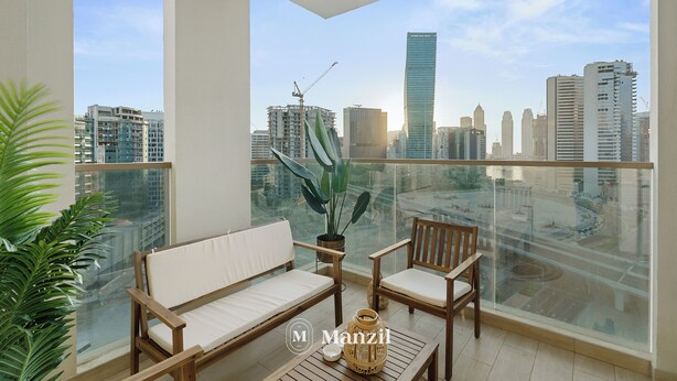 A balcony with a city view, a wooden sofa, a chair, a table, and a potted plant. - 2BR w Canal & Burj View Near Dubai Mall (Dubai)
