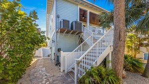 A house with a covered porch, a staircase, and a small garden area with palm trees and shrubs. - Magnolia Cottage: Private Pool, Near Beaches & Pine Ave (Anna Maria)