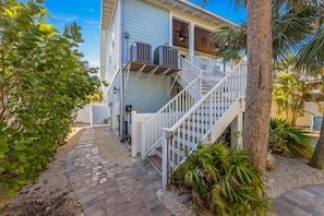 A house with a covered porch, a staircase, and a small garden area with palm trees and shrubs. - Magnolia Cottage: Private Pool, Near Beaches & Pine Ave (Anna Maria)
