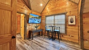 Un accogliente interno di una baita con soffitto in legno, una TV montata a parete, un'area pranzo con tavolo e sedie, e una finestra con persiane.