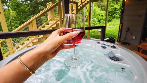 Una mano che tiene un bicchiere di vino rosso sopra una vasca idromassaggio con un pontile di legno e delle scale sullo sfondo.