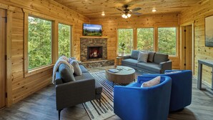 A cozy cabin interior with a stone fireplace, wooden walls, and large windows offering a view of the forest. - Private Indoor Pool Cabin | Game Room + Firepit (Gatlinburg)