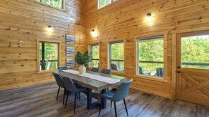 A wooden cabin interior with a dining table, chairs, and large windows offering views of the outdoors. - Private Indoor Pool Cabin | Game Room + Firepit (Gatlinburg)