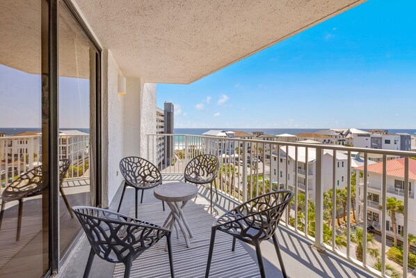 A balcony with a table and chairs, overlooking a cityscape with buildings and palm trees.