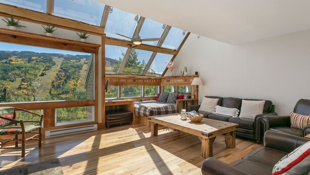 A spacious room with a large skylight, wooden flooring, a sofa, a table, and a view of a forested landscape.