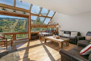 Amplia habitación con un tragaluz grande, suelo de madera, un sofá, una mesa y vistas a un paisaje boscoso.