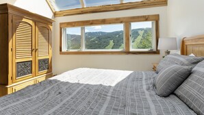 A bedroom with a skylight, a large window with a view of the countryside, a wooden wardrobe, and a bedside table with a lamp.