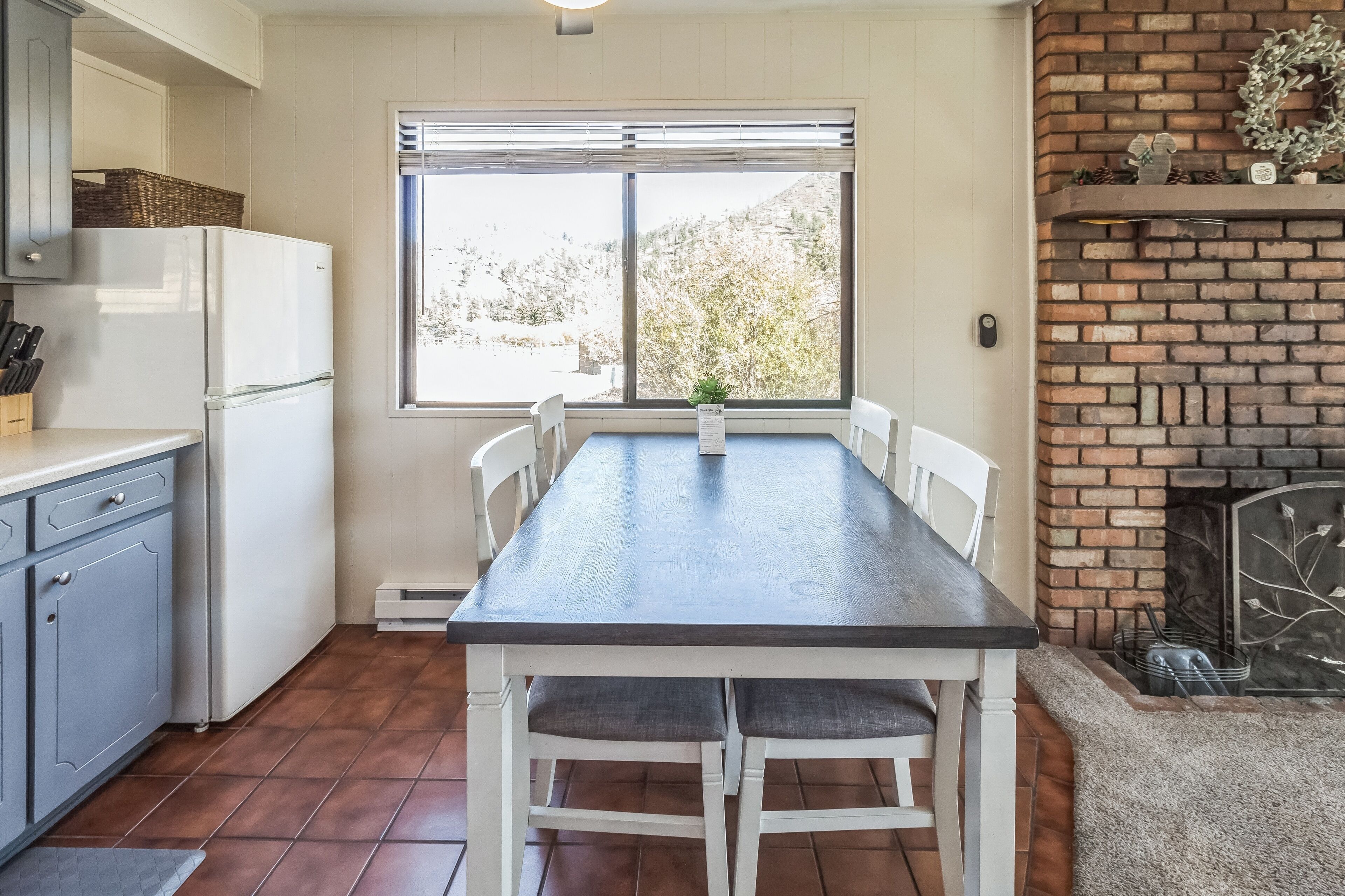 A kitchen with a dining table, white cabinets, a refrigerator, and a brick fireplace.