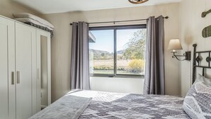 A bedroom with a large bed, a wardrobe, a window with curtains, and a lamp on the wall. - Cozy Cabin Retreat With Scenic Views 2 Bedroom Cabin (Greer)