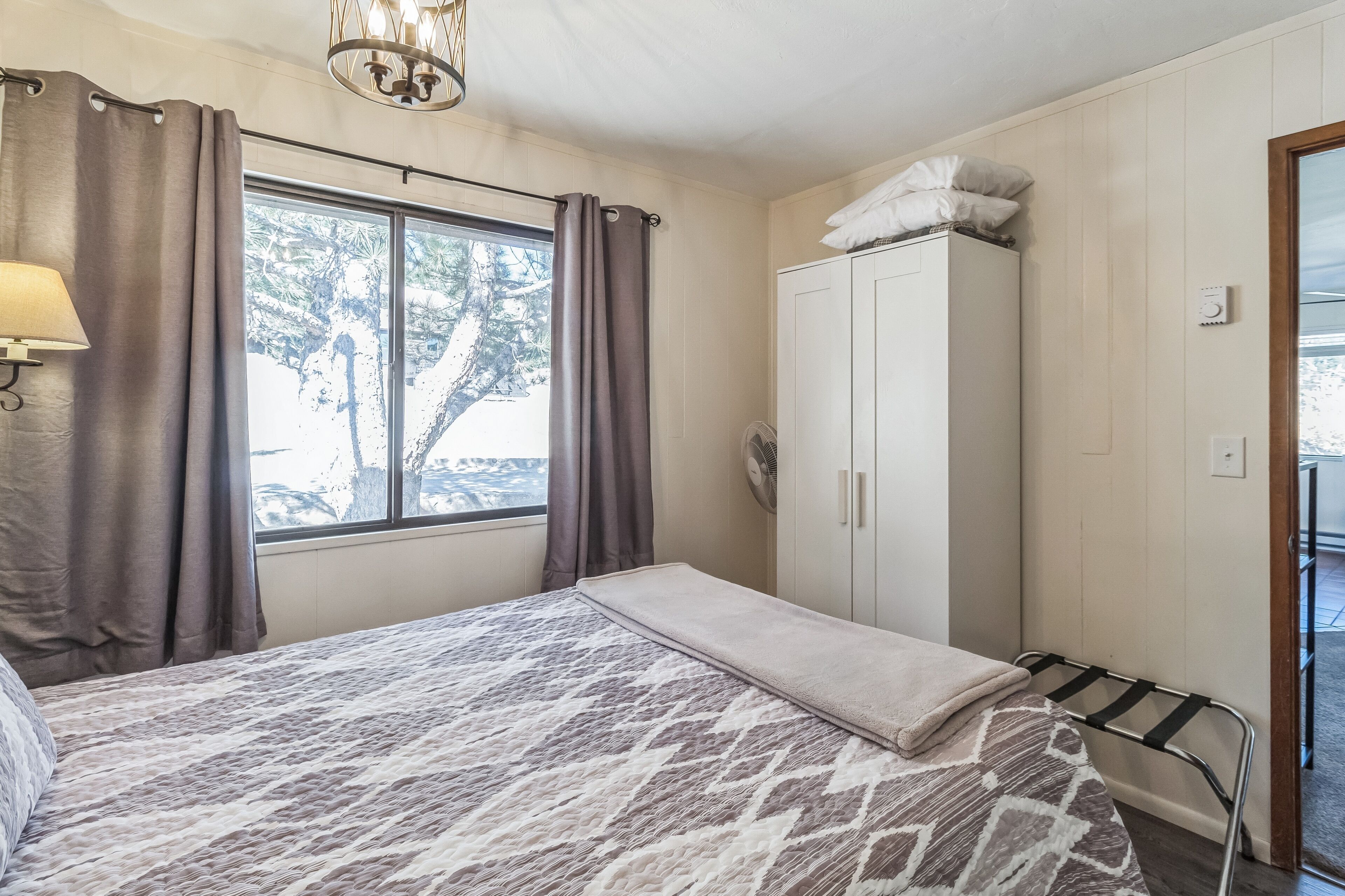 A bedroom with a bed, a wardrobe, a window with curtains, and a lamp.