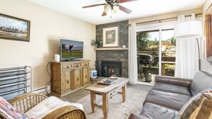 A living room with a fireplace, a television, a ceiling fan, and a view of the outdoors. - E108 W Beaver Creek Blvd 2 Bedroom Condo (Avon)