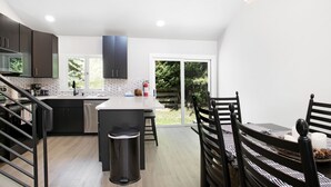 Una cocina moderna con armarios oscuros, encimera blanca y una isla central. Hay una zona de comedor con una mesa y sillas, y vistas a la vegetación exterior.