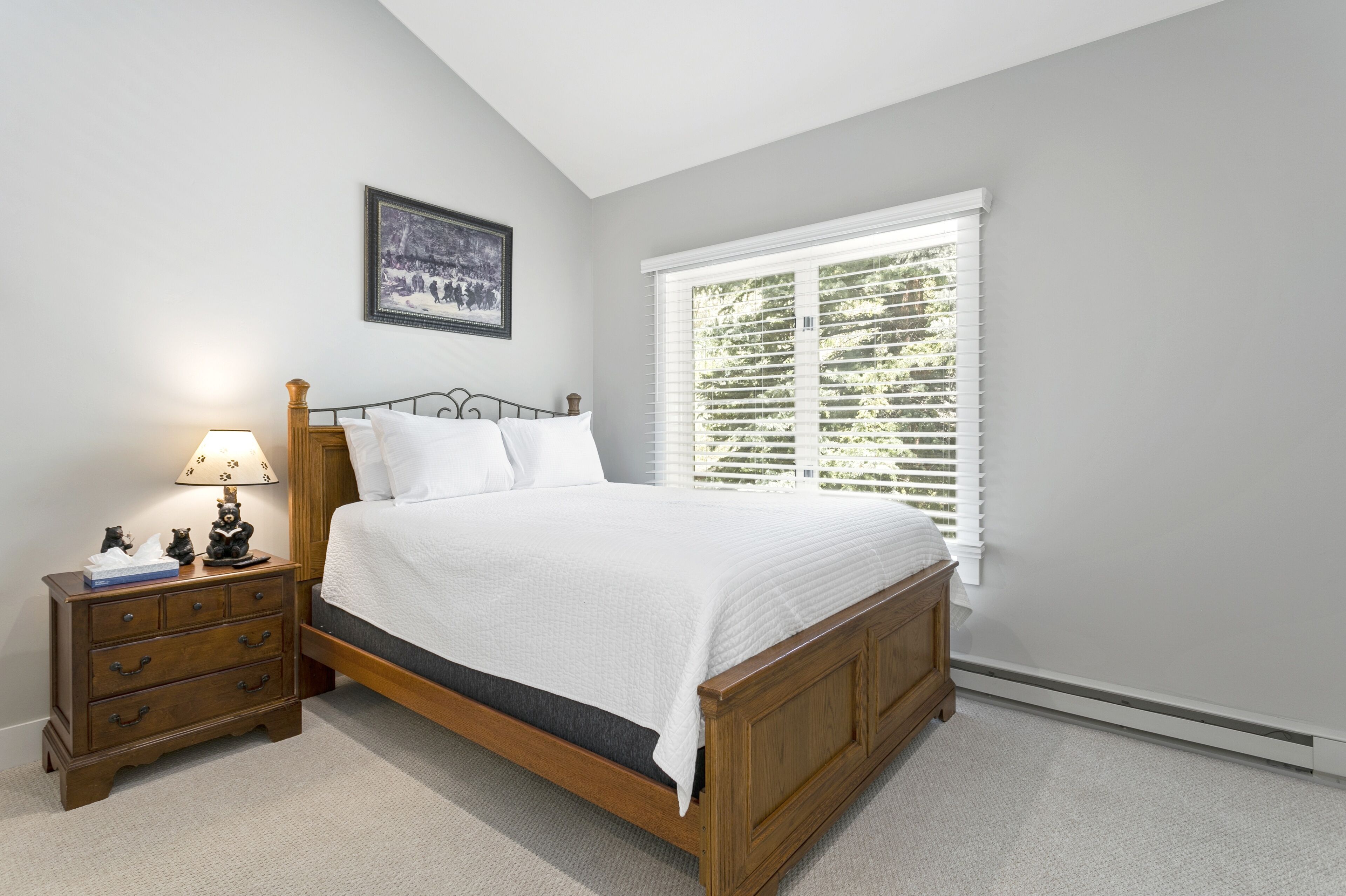 Une chambre à coucher avec un lit en bois, une table de chevet avec des tiroirs, une lampe, un tableau encadré au mur et une fenêtre avec des stores.
