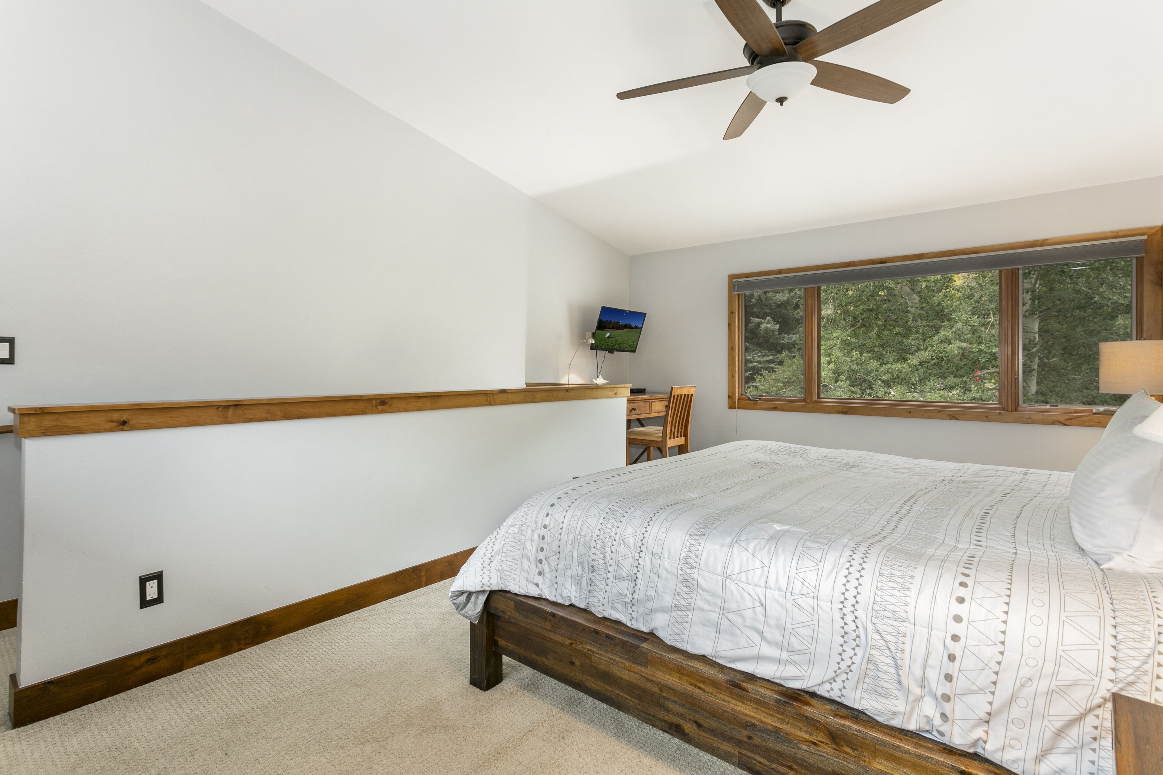 A bedroom with a wooden bed, a ceiling fan, a desk with a computer, and a window with a view of trees.