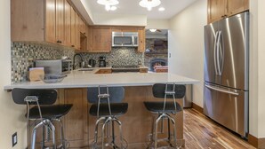 A modern kitchen with wooden cabinets, a stainless steel refrigerator, a microwave, and a tiled backsplash. There are three bar stools and a large island with a sink.