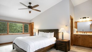 A bedroom with a bed, bedside tables, a ceiling fan, a large window, and a bathroom with a mirror and sink.