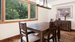 A dining area with a wooden table and chairs, large windows with a view of trees, a chandelier, and a framed picture on the wall.