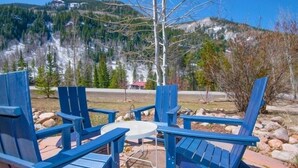 A patio with blue Adirondack chairs and a small round table, overlooking a mountainous landscape with trees and a building in the background. - 4192 Spruce Way 1 Bedroom Condo (Vail)