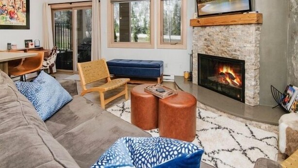 A living room with a stone fireplace, a television mounted above it, and a large window with a view of trees. - 4192 Spruce Way 1 Bedroom Condo (Vail)