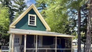 Uma casa pequena, térrea, com telhado verde e varanda coberta.