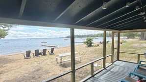 A covered deck with a view of a beach and lake, featuring outdoor seating and a picnic table. - Friendly Inn Limit 5 3 Bedroom Cottage (Sebago)