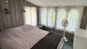 A bedroom with a large bed, a bedside table, a fan, and a window with curtains. - Friendly Inn Limit 5 3 Bedroom Cottage (Sebago)