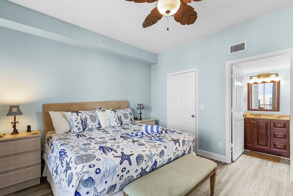 Une chambre à coucher comprenant un lit, des tables de chevet, un banc, un ventilateur de plafond, une porte et une salle de bain avec un meuble-lavabo et un miroir.
